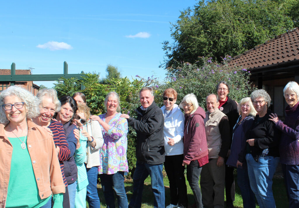 Fourteen Still Green members standing in conga formation in front of shrubbery in the garden of the Milton Keynes Quaker Meeting House