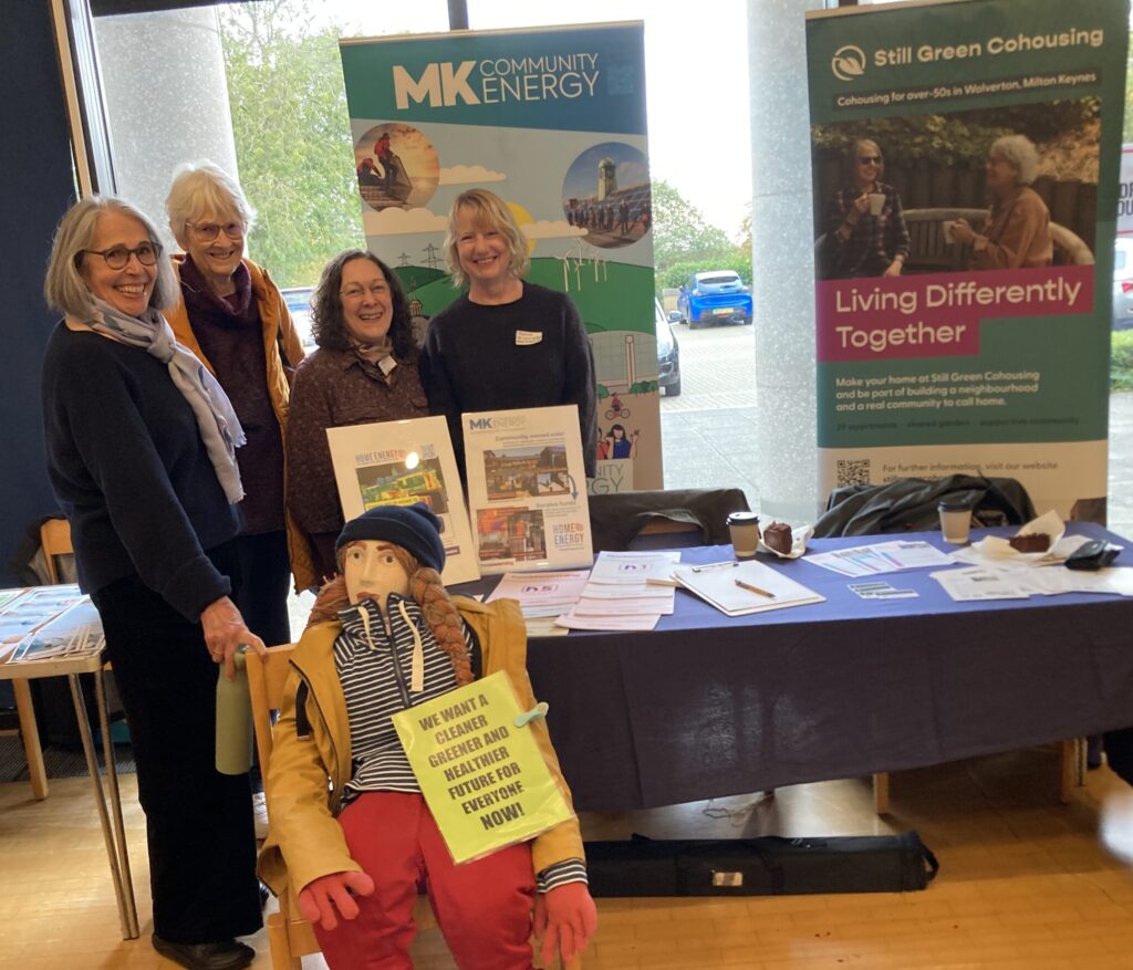 Three members of Still Green are shown standing with a member of MK Community Energy at a stall. A 'Greta Thunberg' scarecrow sits on a chair infront of them, holding a sign about climate action.
